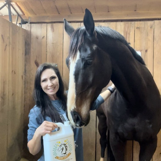 Denise G. in the barn aisle with her off-the-track Thoroughbred and a jug of Camelina Oil